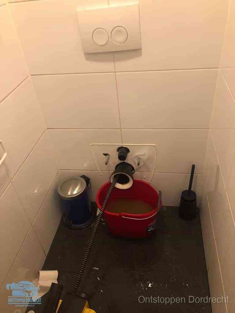 Een ontstopte wand-wc met verwijderde toiletpot waar een ontstoppingsveer in de wanduitgang verdwijnt, het achterhalen waar de verstopping zit in Dordrecht